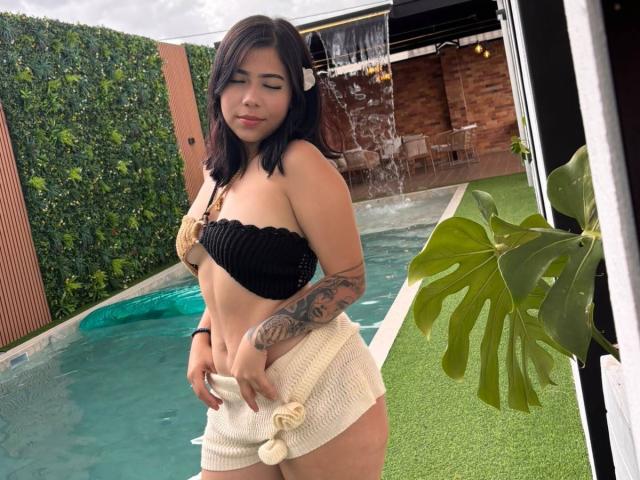 Maddyx69 met kleine borsten, bruine huid, een zwarte top zonder bandjes, witte gebreide shorts, een getatoeëerde arm, zwart haar, staand bij een zwembad met gras, met water dat achter haar valt..