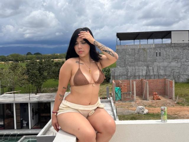 Maddyx69 con grandes senos, piel bronceada, cabello negro, tatuajes, un sujetador de malla marrón, pantalones cortos beige, una mano en la cabeza, sentada en el techo, cielo nublado, edificio de ladrillo gris, césped verde, puede..