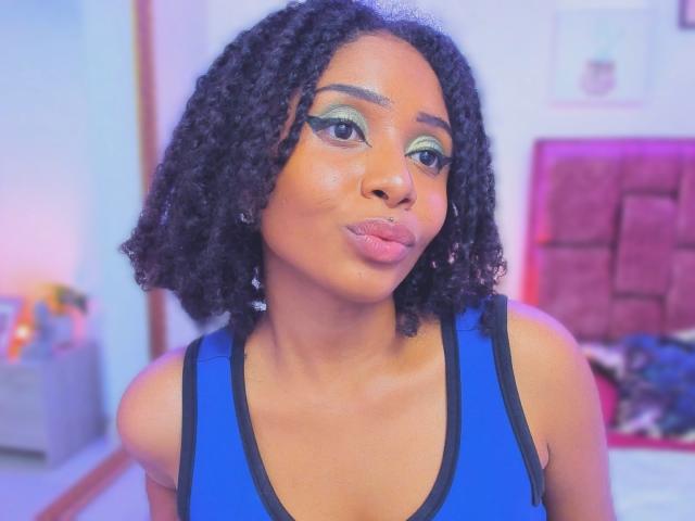 MeganWilliams with dark curly hair, green eyeshadow, black eyeliner, and a nose piercing, pouting her glossy pink lips, wearing a blue tank top with black trim.
