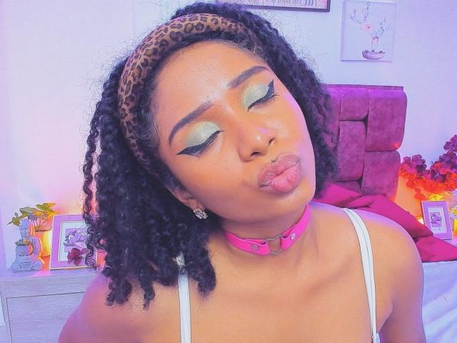 MeganWlliams with puffy lips, pink choker, leopard headband, curly black hair, green eyeshadow, white tank top, nose ring, and pink earrings, puckering up in a selfie.