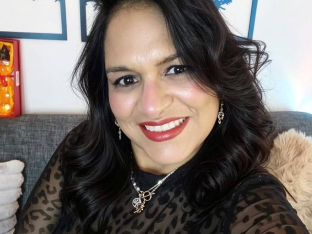 Angela Browm tiene el cabello oscuro y ondulado, lápiz labial rojo, y está sonriendo a la cámara con una piel brillante, vistiendo una blusa de estampado de leopardo y un collar con un dije en forma de corazón..