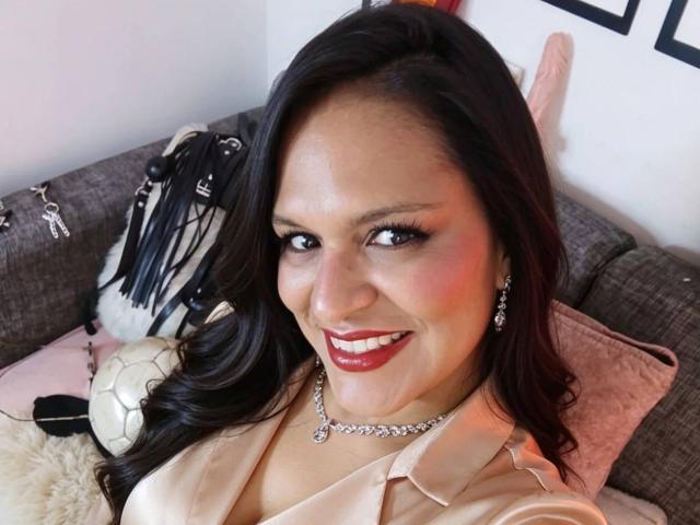 Angela Browm, com cabelo escuro e ondulado, batom vermelho e um sorriso, vestindo uma camisa bege e um colar de diamantes, sentada em um sofá cinza com equipamentos esportivos..