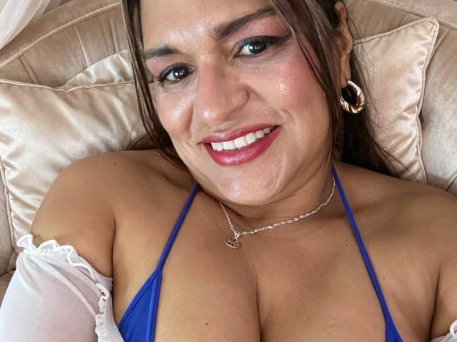 Angela Browm, con cabello castaño, top de bikini azul, mangas blancas, labial rosa brillante, aros grandes, collar, sonriendo a la cámara, mostrando un busto grande, acostada sobre almohadones beige, con el rostro ligeramente sudado..
