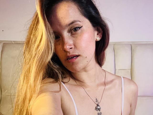 SophiBerta, avec de longs cheveux blonds et bruns, un haut blanc à bretelles spaghetti, un maquillage discret, de petites boucles d'oreilles, un collier avec un croissant de lune, des lèvres légèrement entrouvertes, une peau claire et un visage mignon..