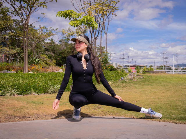AlanaBlake is squatting on a sidewalk, wearing a black bodysuit, black leggings, white sneakers, and a cap, with headphones around her neck, in a sunny park.