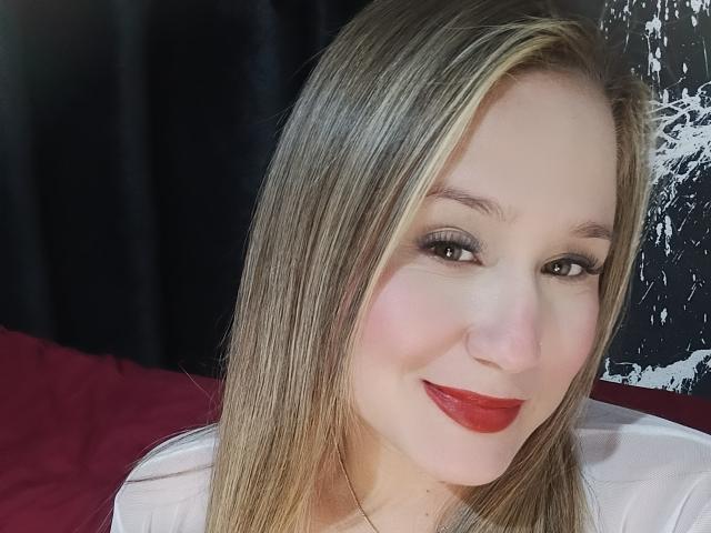 AlexaHotSeins mit blonden Haaren, rotem Lippenstift und Smokey-Eye-Make-up, lächelt in die Kamera. Sie trägt ein weißes Hemd, wobei ihre Brüste teilweise sichtbar sind, und hat ein süßes, sexy Aussehen..
