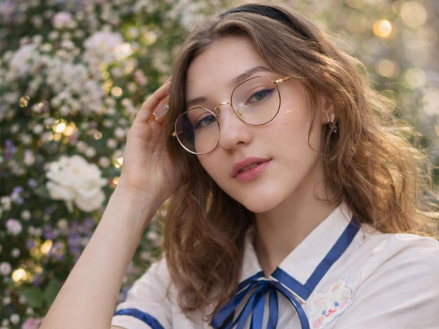 Lenore Schontz, avec ses cheveux bruns ondulés, ses lunettes dorées, sa chemise blanche avec un ruban bleu et des broderies florales, touchant sa tête, une lumière douce, des lèvres roses légèrement entrouvertes, une expression douce..