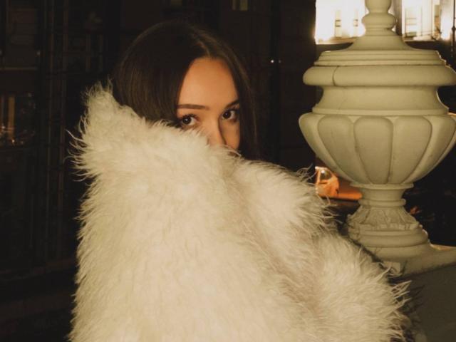 Le visage de Tawnya Kaczmarski est partiellement dissimulé par une fourrure blanche et duveteuse, ses yeux sont tournés vers l'avant, et elle se tient à côté d'une lampe blanche ornée. Elle a les cheveux bruns, et l'arrière-plan est sombre..