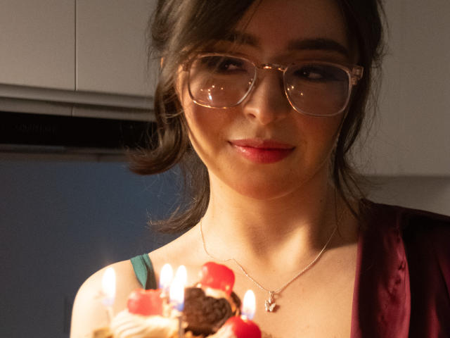 Eva McAllister, con gli occhiali, il rossetto rosso e una collana, tiene in mano una torta con le candeline, sorridendo dolcemente. Il seno è visibile sotto un vestito color borgogna. Capelli castani raccolti in una coda laterale..