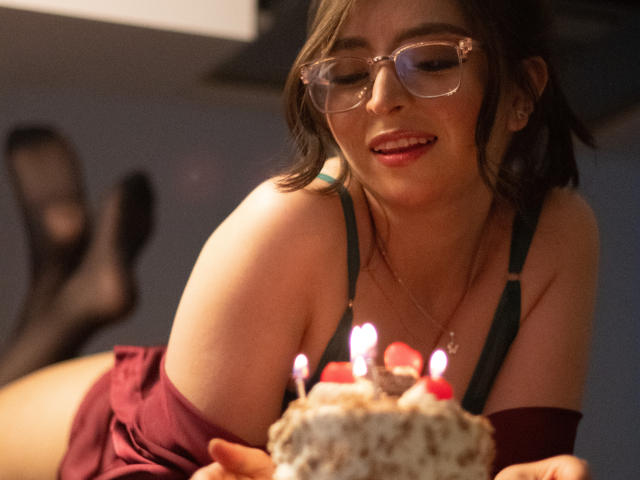 EvaMcAllister, con gli occhiali, sorridente, che tiene una torta con le candeline, indossa un abito verde scuro, con il seno visibile, sdraiata su un divano, gambe con calze nere, rossetto rosso..