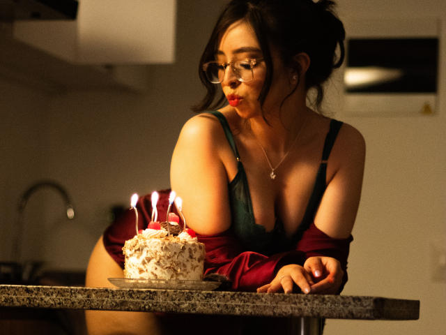 Eva McAllister, con gli occhiali, capelli scuri raccolti in un disordinato chignon, indossa un reggiseno verde e una maglietta rossa a spalle scoperte, è china su una torta con le candeline in una cucina scarsamente illuminata..