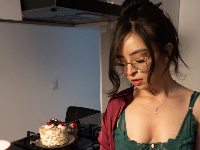 Eva McAllister, con gli occhiali, una camicia rossa e un vestito verde, con un seno di media grandezza, guarda in basso una torta con le ciliegie su un fornello. Ha un viso carino e una collana..