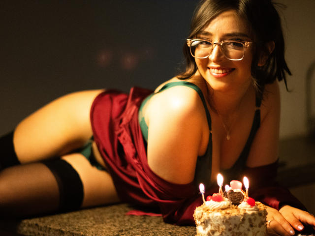 EvaMcAllister, con gli occhiali, sdraiata sul tappeto, sorridente, indossa un vestito rosso, un reggiseno verde, calze nere e biancheria intima verde, accanto a una torta con le candeline, in un ambiente con luce soffusa e calda..