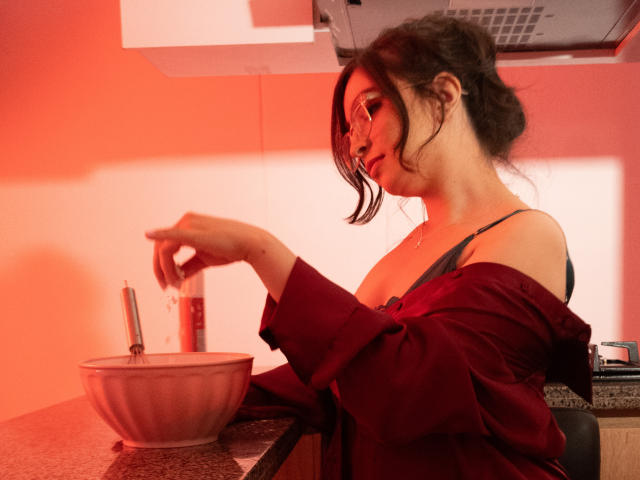 EvaMcAllister con gli occhiali, capelli scuri raccolti in un disordinato chignon, maglietta rossa tolta, reggiseno nero, che versa da una bottiglia in una ciotola rosa, illuminazione rossa, cucina, piano di lavoro maculato..