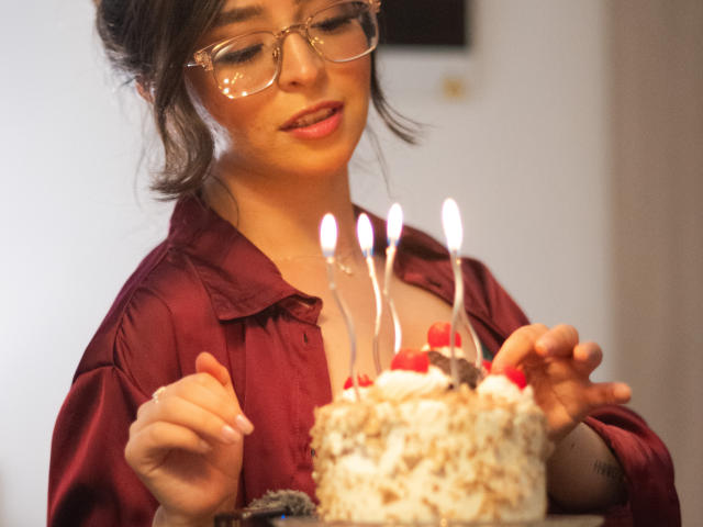 Eva McAllister, con gli occhiali, indossa una camicia rossa, tiene in mano una torta con cinque candeline accese, guarda la torta e sorride. Il seno è leggermente visibile sotto la camicia. Luce soffusa..