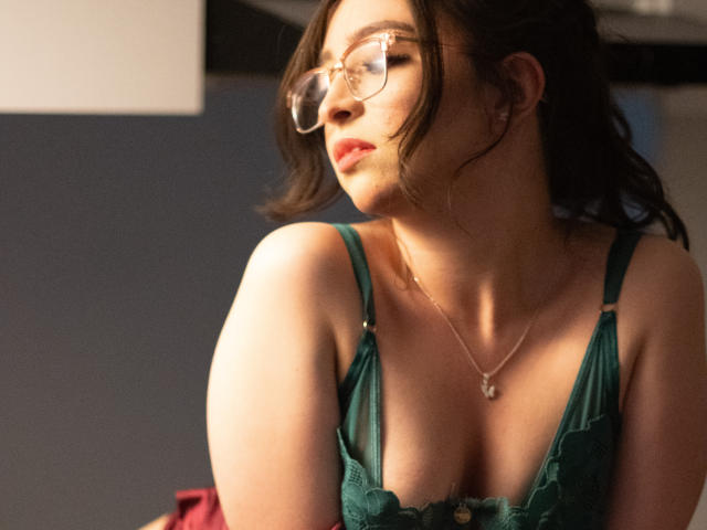 EvaMcAllister con gli occhiali, un reggiseno di pizzo verde e scollatura, che guarda di lato, capelli castano chiaro ondulati, collana dorata, luce solare soffusa, sorriso delicato, leggero rossore, espressione rilassata..