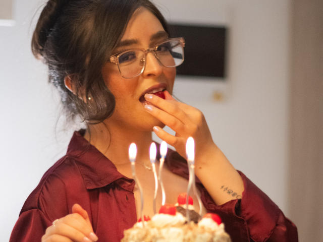 EvaMcAllister con gli occhiali, una maglietta rossa, che mangia una torta con le candeline, una mano vicino alla bocca, un tatuaggio visibile sul polso, capelli scuri raccolti in uno chignon, bocca leggermente aperta..