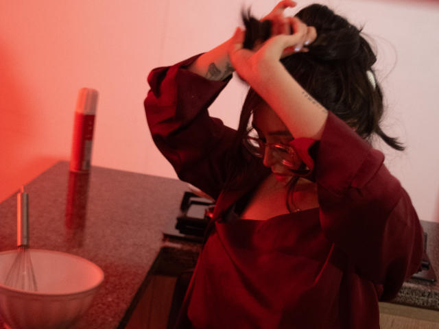 Eva McAllister, con capelli neri, vestito rosso, occhiali, tatuaggio sull'avambraccio sinistro, che usa entrambe le mani per pettinarsi, in piedi in cucina con illuminazione rossa, una ciotola sul bancone..