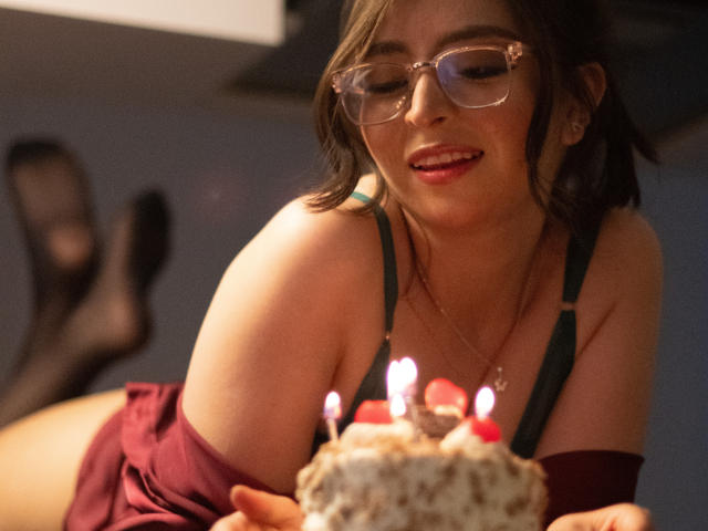 EvaMcAllister con gli occhiali, sorridente, indossa un reggiseno verde e un vestito rosso, tiene in mano una torta con le candeline, mostra i suoi seni, in un ambiente interno accogliente, con le gambe sollevate..