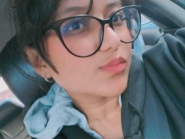 AzulMoon with black glasses, light blue eyeshadow, and pink lips, wearing a grey hoodie and seatbelt, looking slightly to the side in a car.