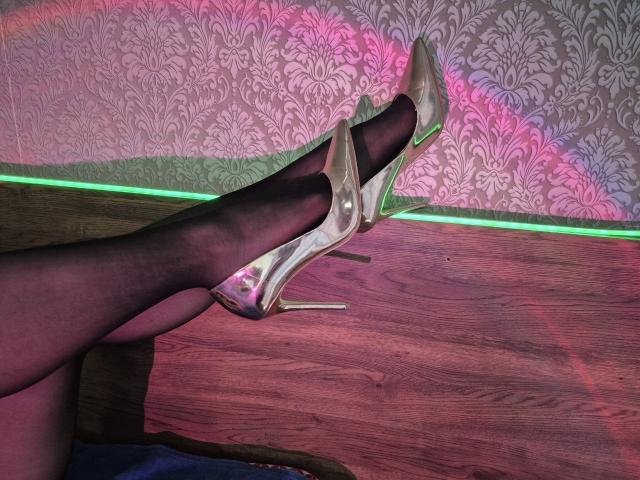 Milagres' legs in sheer black stockings with white stiletto heels, crossed at the ankle, resting on a wooden surface, with pink and green LED lights.