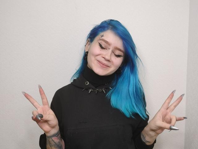 BrittanieMcclaran with blue hair, black hoodie, and tattoos, smiling, showing dual peace signs, has star tattoos on cheek, long nails, and a nose ring.