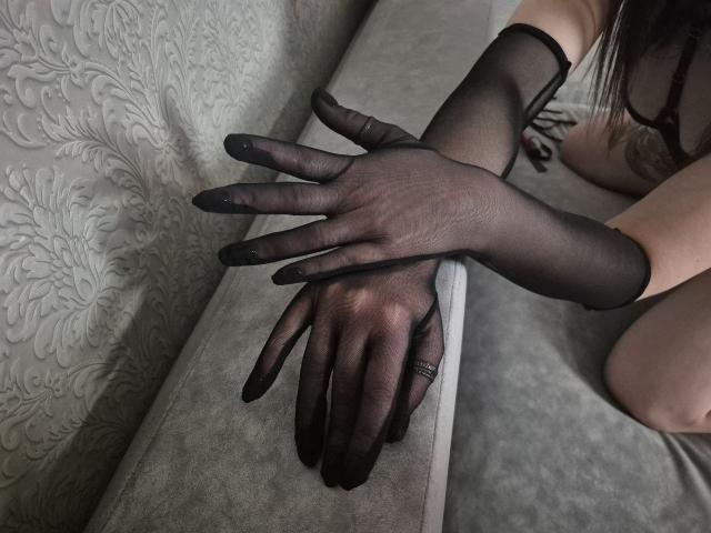 Milagres' hands in sheer black gloves, fingers spread, resting on a gray sofa. Her ass is partially visible in the background, sitting with legs apart.