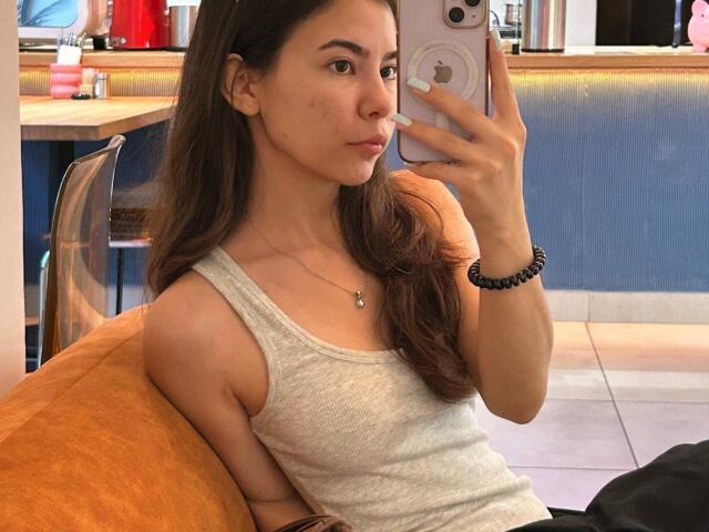 EmeraldCrivaro with long brown hair, wearing a grey tank top, sitting on an orange couch, taking a mirror selfie with a white iPhone, showing her small tits and pale skin.