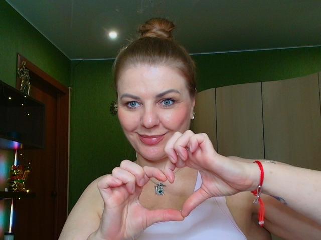 VeronikaGoldy con i capelli biondi raccolti in uno chignon, occhi azzurri, che sorride, indossa una canotta bianca, forma un cuore con le mani, braccialetto rosso, parete verde sullo sfondo..