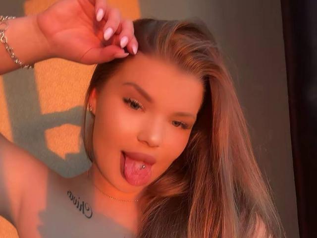 NicoleFlameX heeft lang, recht bruin haar, een doorboorde tong en een tatoeage op haar linker schouder. Ze steekt haar tong uit en heft haar hand op. Warm, oranje licht..