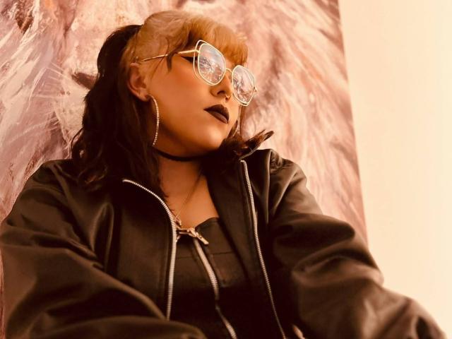 YamiiRyuu com cabelo laranja, óculos, batom preto, brincos de argola, colar, jaqueta preta e blusa preta, olhando para cima, exibindo uma pequena e fofa parte traseira. Ambiente urbano, acolhedor e iluminado..