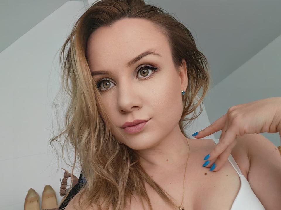 HeartlessQueenn with blonde hair, blue nails, and white top, pointing to her chin, has green eyes, pink lips, and a small mole on her chest.