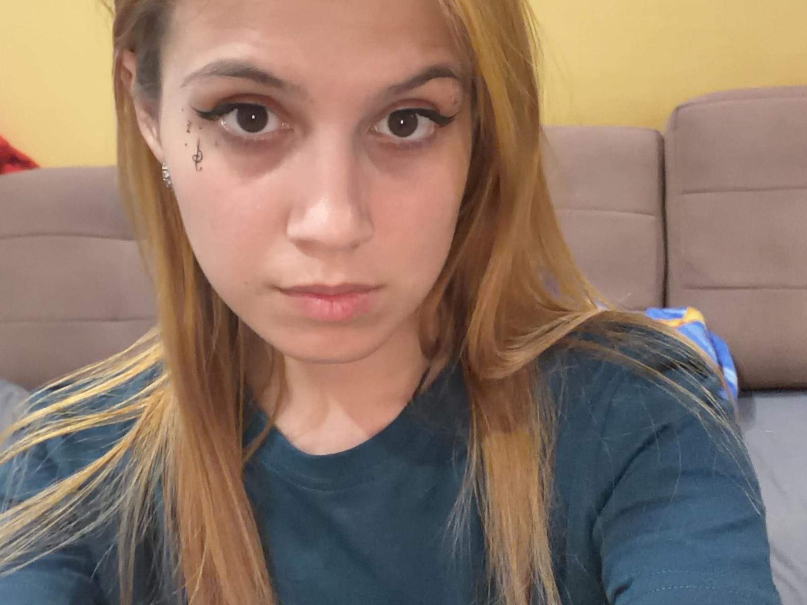 EsterLagreca heeft een lichte huid, bruine ogen en lang blond haar, draagt een turquoise shirt, heeft een kleine zwarte tatoeage vlakbij haar rechteroog en een serieuze uitdrukking op haar gezicht..