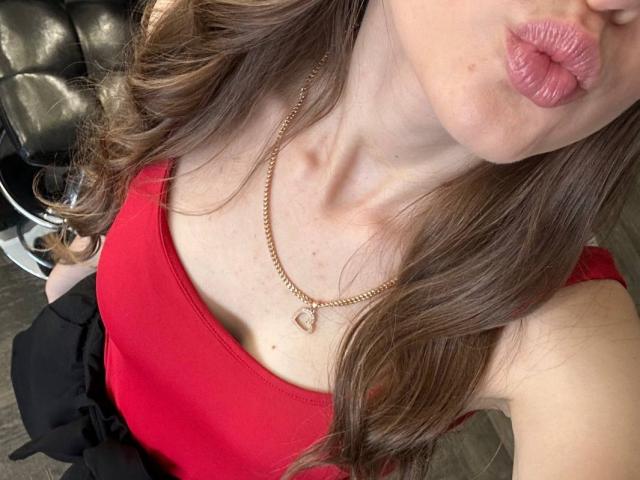 A blusa vermelha de Diantrisy, que mostra seus pequenos seios e clavícula, com um colar dourado e lábios rosados. Cabelo castanho ondulado, calças pretas. Parte superior do corpo, levemente inclinada..