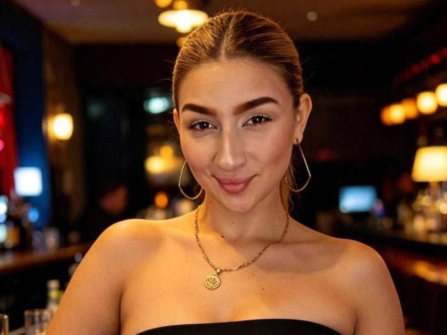 Abby, una mujer con cabello castaño recogido en una coleta, que lleva grandes aros y un top negro sin tirantes, sonriendo sutilmente, con un collar dorado, iluminación cálida, y un fondo de bar oscuro..