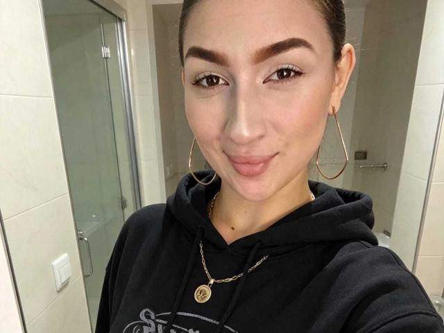 AbbyMilers tem a pele de cor marrom clara, sobrancelhas marrons escuras, e está usando brincos grandes de argola dourados, um moletom preto e um colar dourado com um pingente circular, sorrindo..