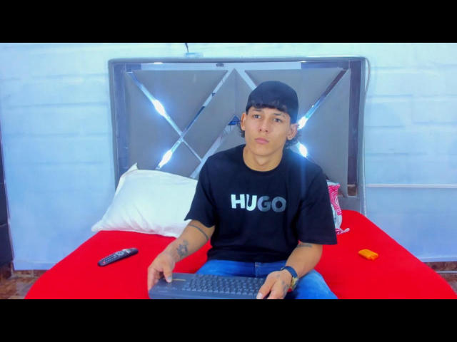 TemptKing, con cabello corto y castaño, gorra negra, camiseta negra de HUO, tatuaje en el brazo derecho, sentado en una cama roja, con un teclado negro, un control remoto, una lata y un anillo naranja frente a él..