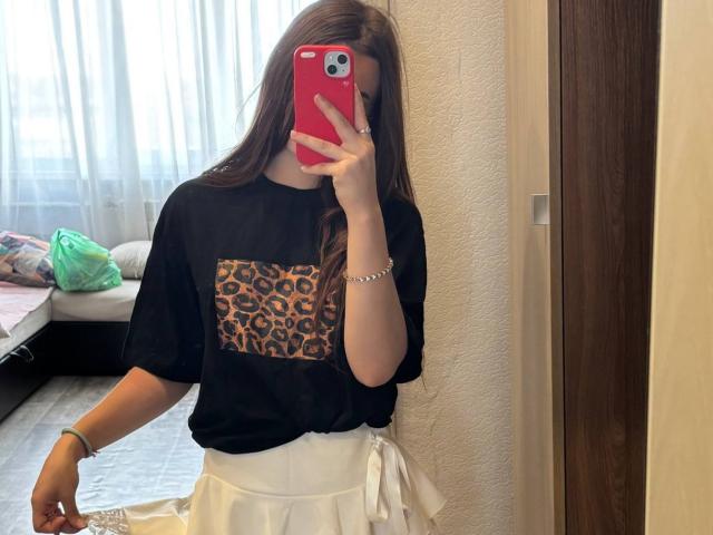 Kimberly Zampieri mit langen braunen Haaren, einem schwarzen Oberteil mit Leopardenmuster, einem weißen Rock, die ein rotes Telefon hält, ein Selfie vor einem Spiegel, eine weiße Wand, eine Holztür, ein weißer Boden und eine grüne Tasche im Hintergrund..