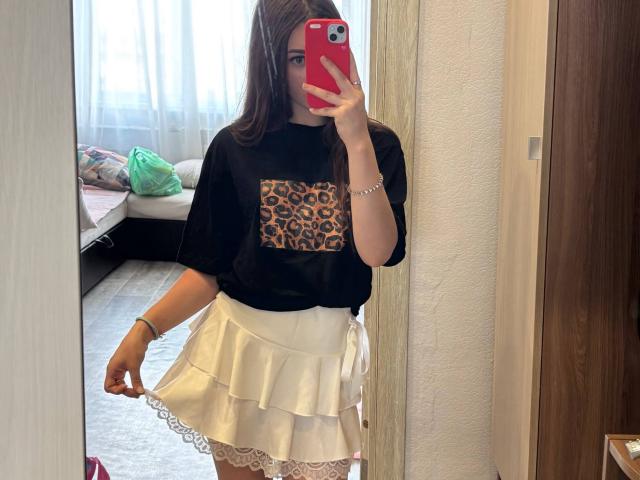 KimberlyZampieri in een spiegelzelfportret, gekleed in een zwart shirt met luipaardprint, een witte rok met ruches en kant, met een rode telefoon in haar hand, lang bruin haar, een witte muur, een houten deur..