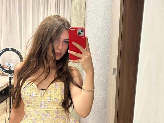 Kimberly Zampieri, met lang bruin haar, draagt een gele jurk met bloemenprint, houdt een rode telefoon vast en maakt een spiegelzelfportret, waarbij haar borsten zichtbaar zijn, in een kamer met een ringlamp..