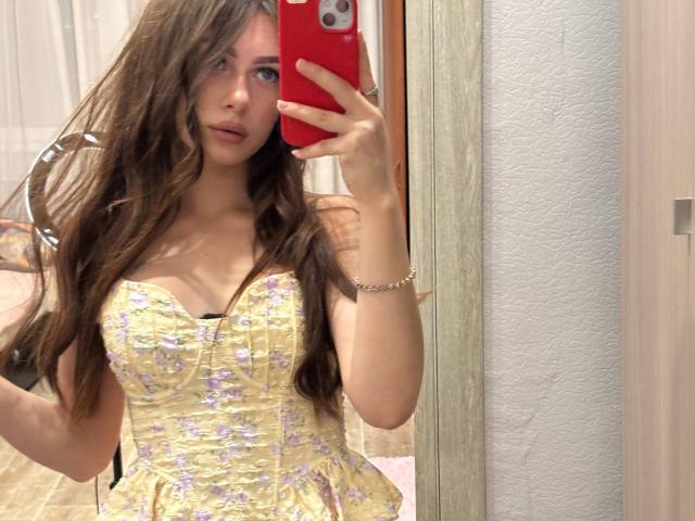 Kimberly Zampieri, met lang bruin haar, draagt een gele jurk met bloemenprint, houdt een rode telefoon vast en maakt een spiegelzelfportret, waarbij haar kleine borsten en lichte decolleté zichtbaar zijn..