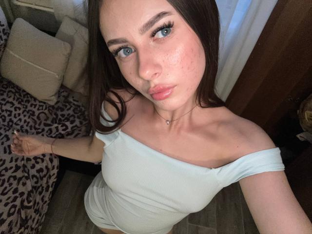 Kimberly Zampieri mit blauen Augen, die ein weißes Top mit einem Schulterauschnitt trägt, wobei ihre Brüste sichtbar sind, mit braunen Haaren und rosafarbenen Lippen, in einem Schlafzimmer mit Bettwäsche mit Leopardenmuster..