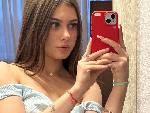 Kimberly Zampieri, con lunghi capelli castani, pelle chiara e labbra rosa, sta scattando un selfie con un telefono rosso, indossando una maglietta azzurra con una spalla scoperta, che mette in mostra piccoli seni e tatuaggi..