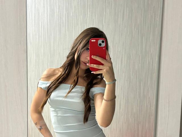 Kimberly Zampieri, met lang bruin haar, draagt een witte top met een open schouder, houdt een rode telefoon vast, bedekt haar gezicht, en toont haar borsten en een kleine tatoeage op haar rechterpols..
