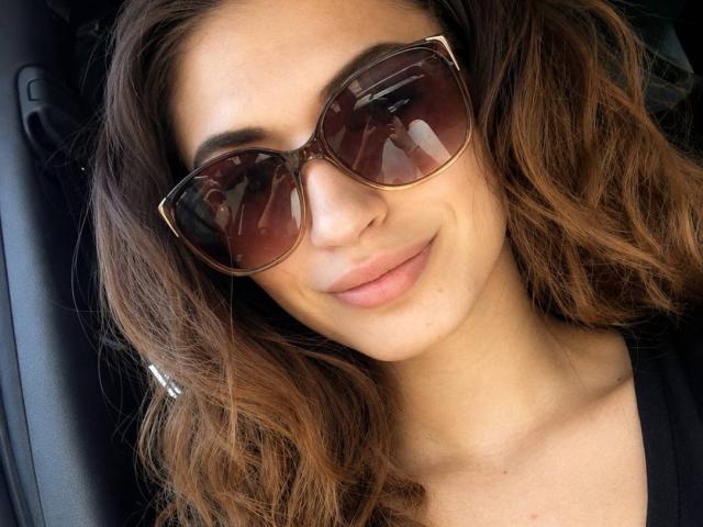 AyakaSweety tiene el cabello castaño ondulado, usa gafas de sol marrones grandes y sonríe ligeramente con labios rosados. Lleva una blusa negra y muestra una cara linda y relajada..