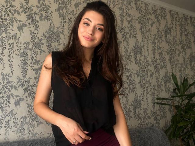AyakaSweety con lunghi capelli castani, indossa una canotta nera, mostrando il seno, in piedi, sorridente, con la mano destra sul ventre, gonna color borgogna, pianta verde a destra..