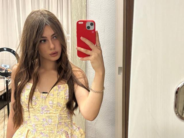 Kimberly Zampieri mit langen, braunen Haaren, trägt ein gelbes Kleid mit Blumenmuster, hält ein rotes Telefon und macht ein Selfie im Spiegel, wobei ihre Brüste im Spiegelbild zu sehen sind..