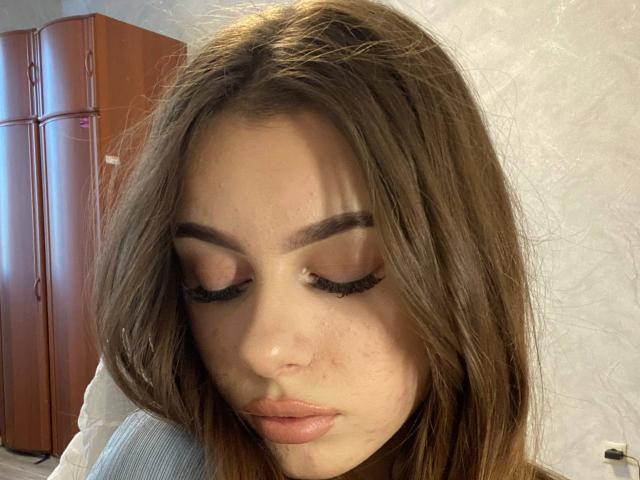 Het gezicht van Kimberly Zampieri, met bruin haar, gesloten ogen en lange zwarte wimpers, met sproeten op haar neus, en ze draagt een lichtgrijze blouse, in een licht verduisterde kamer..