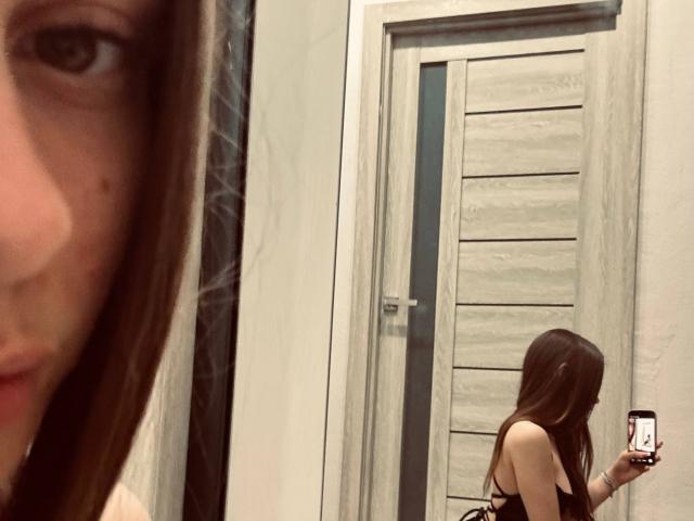 KimberlyZampieri's reflectie toont haar met lang bruin haar, kleine borsten en een telefoon in haar rechterhand, staand voor een houten deur in een spiegelzelfportret..