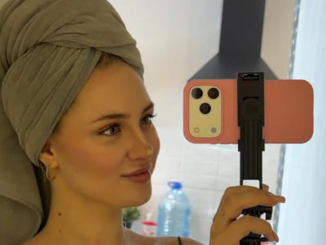 NancyBijou com uma toalha na cabeça, tirando uma selfie com um iPhone rosa, segurando um tripé preto, pequeno piercing no nariz, sorriso discreto, fundo branco..